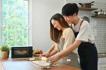 Fototapeta na wymiar Attractive man and woman in casual outfits kneading dough with a rolling pin on wooden table in kitchen interior