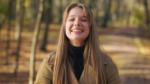 Front view of attractive, young lady standing in park in autumn. Happy, cheerful woman looking at camera, smiling, showing hurray. Concept of happiness and sincere emotions.
