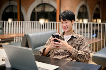 Relaxed Asian male freelancer or college student using his smartphone, remote working at cafe