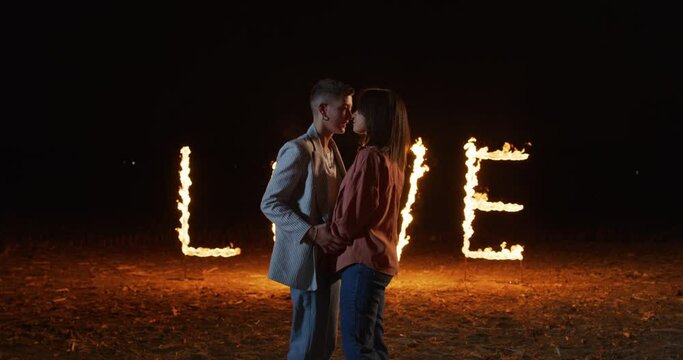 Portrait of Young Homosexual Gay Female Couple kissing With the background of the Word LOVE composed of glowing sparkling pyrotechnic fire flame letters. Lesbian Lovers Celebrating their Relationship 