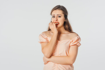 Fototapeta premium Anxious young woman bites fingernails, looks nervous, scared of smth, stands over white background. Amazed horrified female keeps hand on mouth