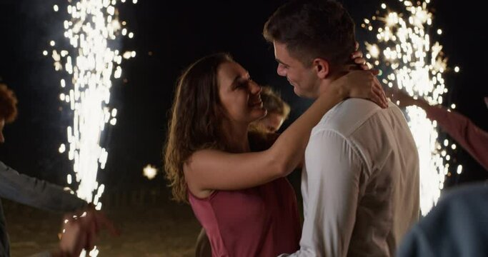 Cinematic Shot of Young Romantic Couple in Love Kissing Surrounded by Friends Dancing with Sparklers During Engagement Party Outdoors at Night. Happy Man and Woman Starting their Life Together 