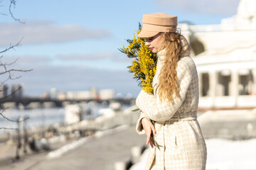 A young woman stands with a bouquet of yellow mimosa and sniffs the flowers. The concept of the...