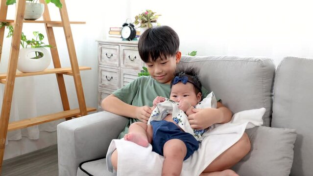 Asian elder brother and newborn baby sit in living room together, adorable infant sitting on boy lap with trust and comfortable. kid hold toddler in embrace with love and care. happy family concept.
