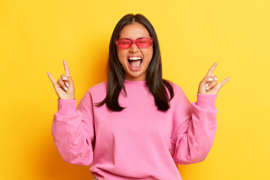 Horizontal shot of happy crazy Asian woman makes rock and roll gesture heavy metal sign has fun dressed in pink casual jumper isolated over yellow background