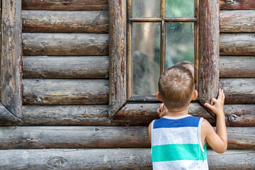 Excited preschooler boy looks into window of ancient log house. Curious child looks at reflection...
