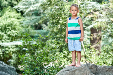 Naklejka na ściany i meble Thrilled boy stands on top of big rock against blurry green trees. Excited child in striped t-shirt enjoys spending summer holidays in park