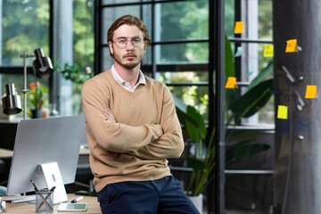 Portrait of a young handsome man freelancer, programmer, He sits on the desk in the office, crosses...