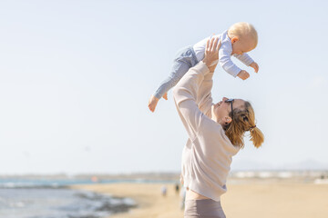 Fototapeta na wymiar Mother enjoying summer vacations holding, playing and lifting his infant baby boy son high in the air on sandy beach on Lanzarote island, Spain. Family travel and vacations concept