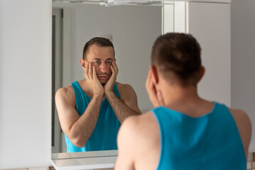 Sleepy tired man in bathroom looks at himself in mirror. Male touching his beard and face. Morning routine