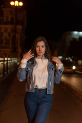 Portrait of girl student walking on night street. Stylish attractive young woman in denim jacket, evening city