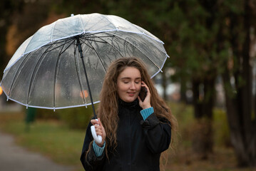 Smiling Young woman talking on phone in autumn park with transparent umbrella. Girl walking in the park and chat