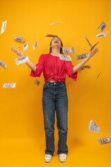 Happy laughing girl with money around boasting wealthy life. Indoor studio shot on orange or yellow background. Vertical frame.