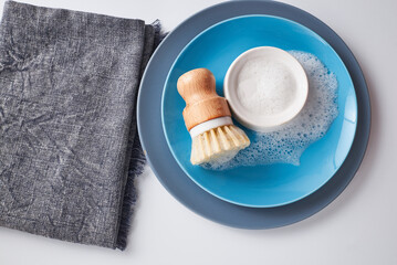 Dishwashing soap on pile of plates with foamy kitchen cleaning sisal bamboo round dish brush. Minimalist white wood background.
