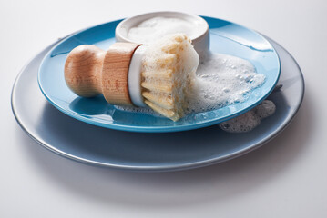 Dishwashing soap on pile of plates with foamy kitchen cleaning sisal bamboo round dish brush. Minimalist white wood background.