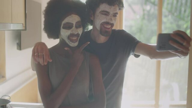 Medium shot of young diverse couple with cosmetic masks on faces posing for smartphone camera while taking selfie during skincare routine at home