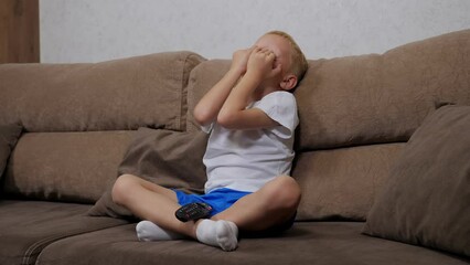 Close-up of a sad little blond boy sitting on the sofa at home, holding a remote control. The child is punished and does not watch TV.