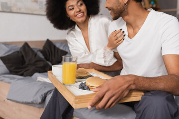 Fototapeta na wymiar smiling african american woman touching boyfriend holding tray with delicious pancakes and orange juice in bedroom