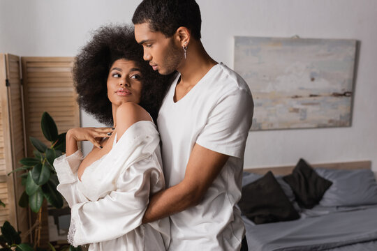 young african american man hugging sexy woman in white silk robe in bedroom