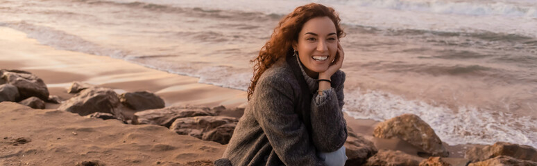 Cheerful young woman looking away while sitting on beach during sunset, banner.