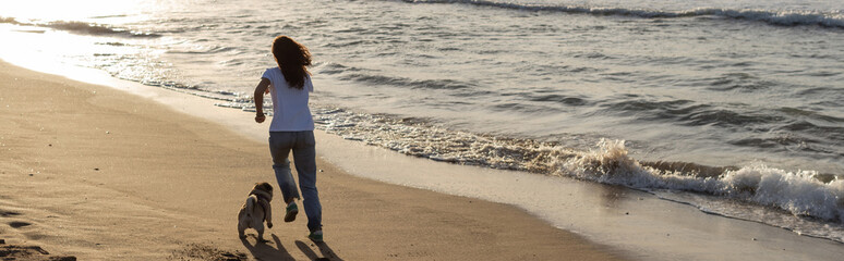 back view of woman running with pug dog on sandy beach during sunset, banner.