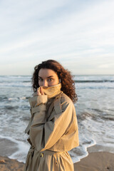 curly woman in wired earphones adjusting collar of beige trench coat and looking at camera near sea in Barcelona.