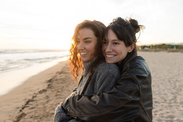 Smiling woman hugging friend on blurred beach during sunset.