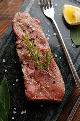 The row ribeye steak with black pepper,  rosemary branch, at the stone modern plate at wooden background with steel fork, knife