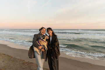 Happy friends holding smartphone and pug dog on beach in Spain.