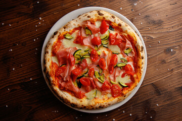 The top view of delicious Italian pizza with tomato cherry, ham, zucchini, mozzarella on the wooden background 