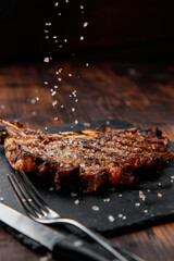 top view of delicious ribeye beef steak on the ribbon on the black stone plate with coarse sea salt falling on it with steel fork and knife in the wooden backgrounds 