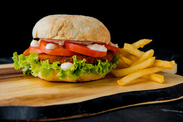 Classic hamburger with pork cutlet, tomato, salad leaves, white sauce with french fries on the wooden plate on the dark background   