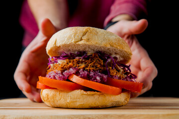 hamburger with beef cutlet, tomato, red marinated onion with hands holding on the wooden plate on the dark background