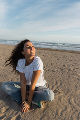 Fototapeta na wymiar dreamy woman sitting in blue jeans and white t-shirt sitting on sandy beach in Spain.