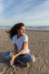 positive woman sitting in blue jeans and white t-shirt sitting on sandy beach in Spain.