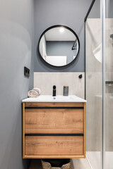 Bathroom with a washbasin on a floating vanity. On a gray wall is a round mirror in a black metal frame. Bathroom is filled with a fashionable design solution, pleasant and comfortable little things.