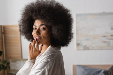 Fototapeta na wymiar seductive african american woman in white silk robe touching lip and smiling at camera at home