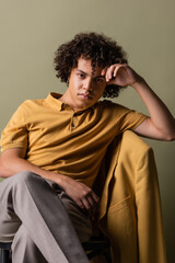 trendy african american man in yellow polo shirt sitting with crossed legs and hand near head on grey background