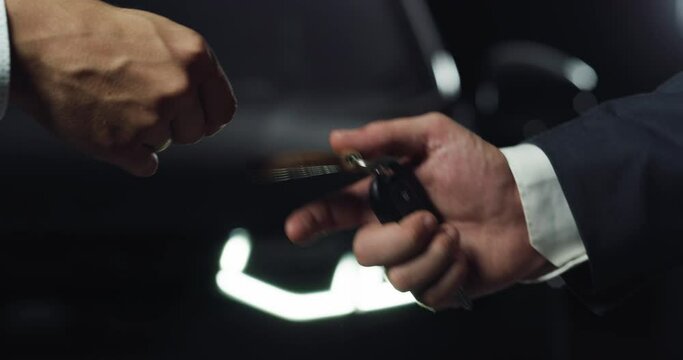 Close Up of Two Male Hands Exchanging a Car Key During a Successful Sale, with a Fancy Anonymous Car in the Background. Businessmen Shaking Hands and Concluding a Beneficial Deal by Buying a Vehicle