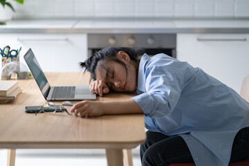 Tired exhausted Asian woman sleeping in kitchen with head on desk near laptop after using social...