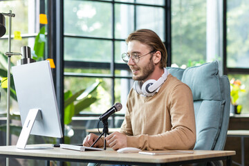 Young handsome male blogger sitting at the desk in the office at the computer in front of the...