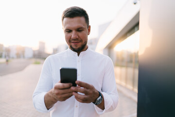 Smiling man looking at screen of smartphone while standing on street