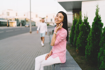 Cheerful Asian woman talking on smartphone sitting on bench on street