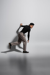 full length of young african american man in black turtleneck and trendy beret looking at camera while walking on grey background