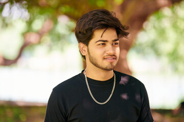 Attractive young indian man portrait in black t shirt and silver neck chain on outdoor green park...