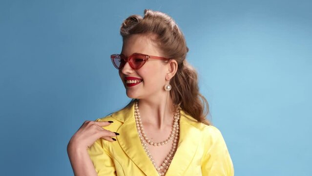 Flirty look. Stylish, beautiful blonde girl posing in trendy sunglasses over blue studio background. Femininity. Concept of retro fashion, beauty, attraction, 50s, 60s. Pin-up style. Vintage