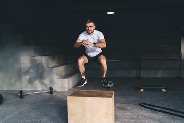 Young man doing squats in air on wooden box in gym