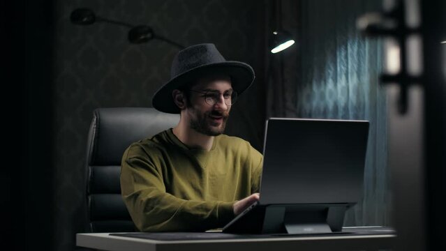 Young man thinking have an ideas sitting at desk in living room.Inspiration guy using laptop work remotely planning job.Beard man wear hat with eyeglasses finding solution of problem indoors.