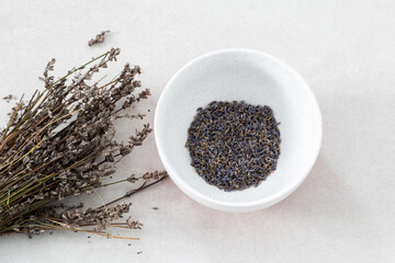 Selective focus flat lay side lit view of bunch of lavender twigs next to flowers in lightly speckled white bowl on ceramic background