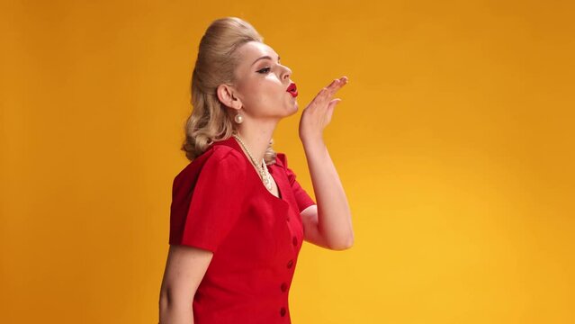 Sending kisses. Beautiful blonde girl with red lips make-up, stylish hairstyle posing over yellow studio background. Concept of retro fashion, beauty, attraction, 50s, 60s. Pin-up style. Vintage